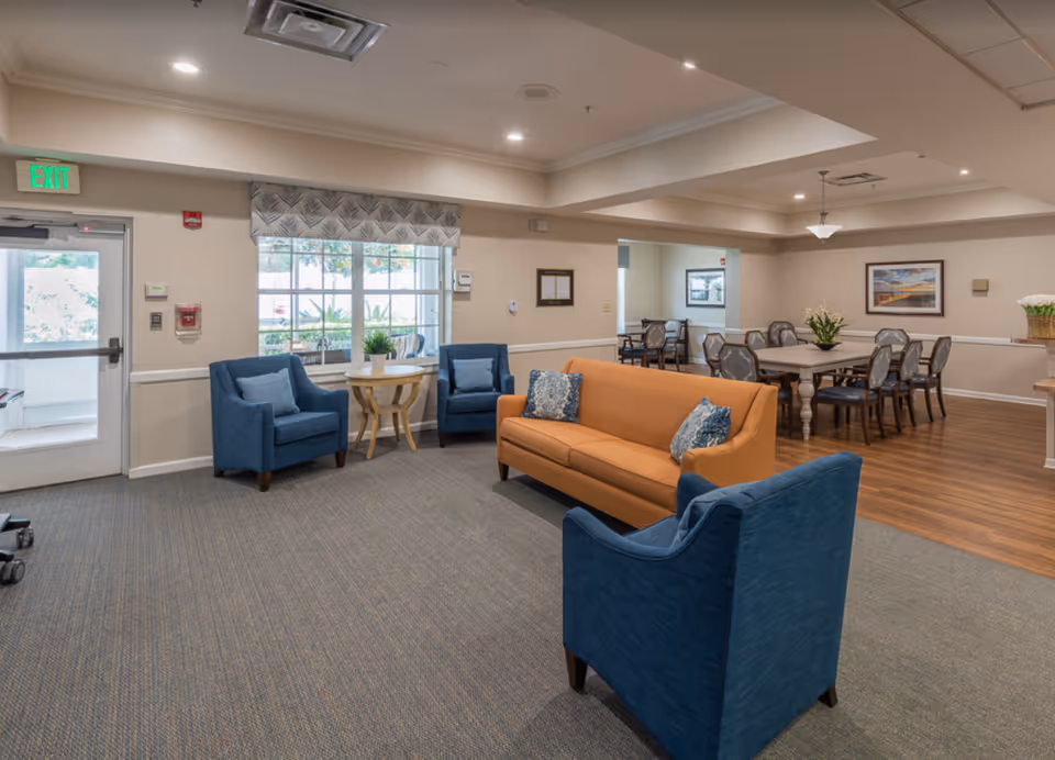 A bright senior living common area with blue armchairs, an orange sofa, and a dining table in the background.