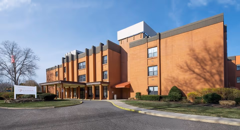 Exterior front of a multi-story orange-brick rehabilitation and nursing building with a covered entrance, driveway, landscaped lawn and a sign reading Luxor.