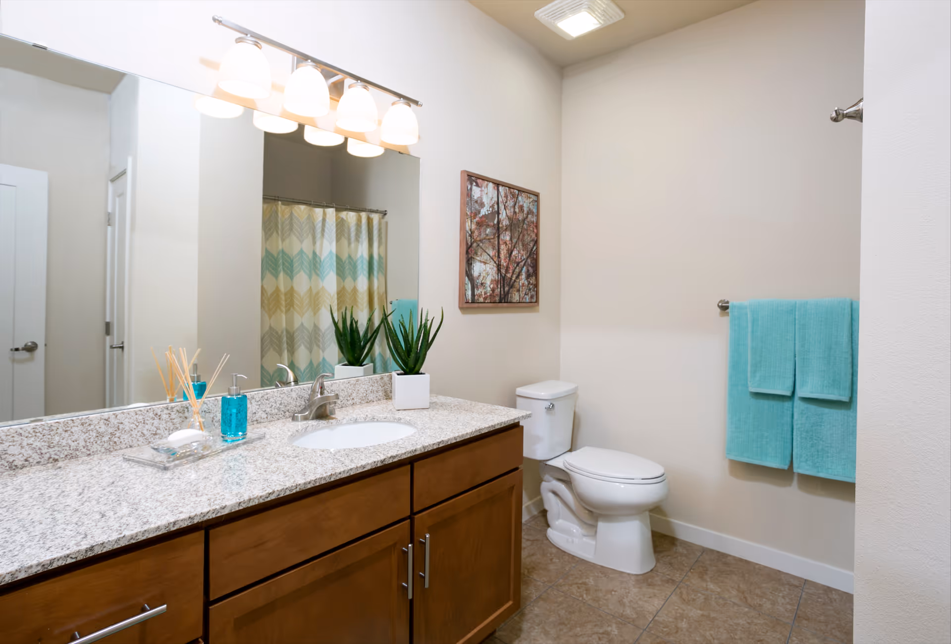 A clean bathroom with a granite countertop vanity featuring a sink, soap dispenser, and a small potted plant. Above the vanity is a large mirror with four light fixtures. To the right is a white toilet, a towel rack with two turquoise towels, and a framed picture on the wall. In the background, a shower curtain with a chevron pattern in shades of beige, gray, and turquoise is visible.