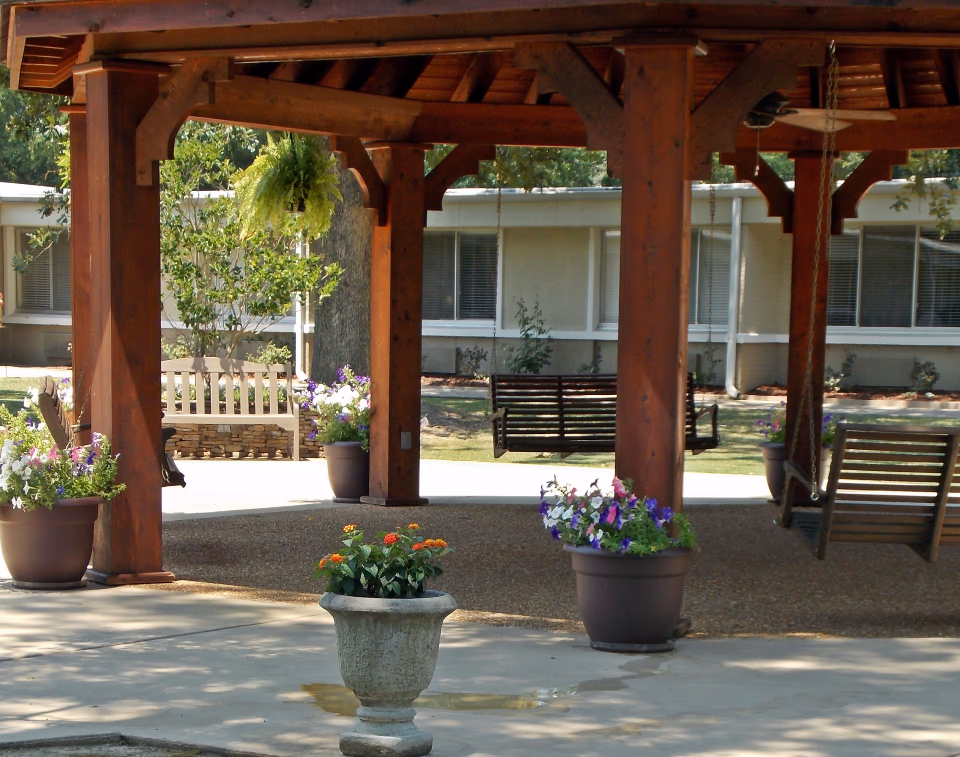 Outdoor covered seating area with wooden beams and hanging swings, surrounded by potted flowers and greenery, with a building visible in the background.