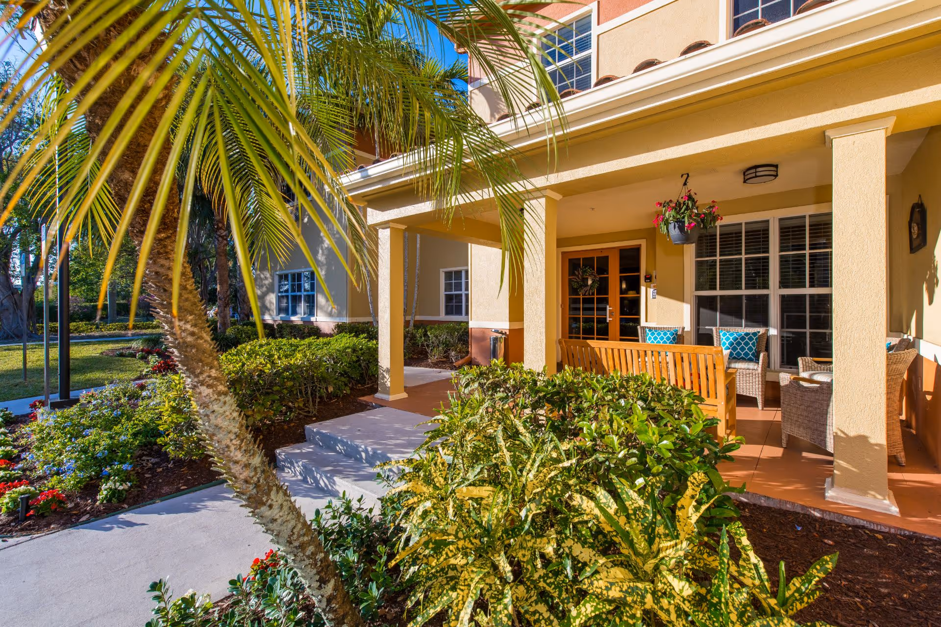 Outdoor patio area at Sonata Delray Beach with a wooden bench, wicker chairs with blue patterned cushions, hanging flower pot, and lush green plants and palm trees surrounding the walkway and porch.