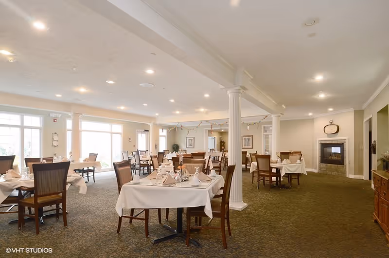 A spacious dining room with multiple tables covered in white tablecloths, each set with napkins, glasses, and silverware. The room features carpeted floors, large windows letting in natural light, white columns, and a fireplace with a clock above it on the far wall.