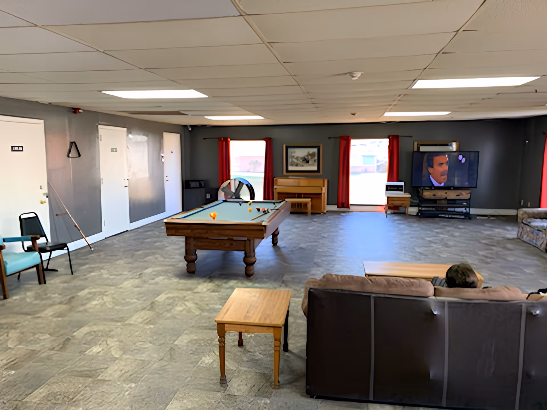 Large communal living/recreation room with a pool table, couches, a TV, and a piano under red-curtained windows.