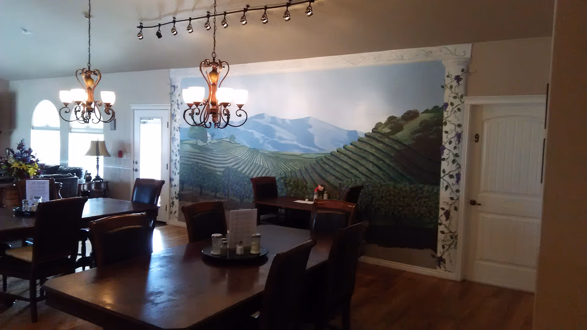 Dining area with wooden tables and chairs, two ornate chandeliers hanging from the ceiling, a mural of vineyard hills on the wall, and a door labeled with the number 9.