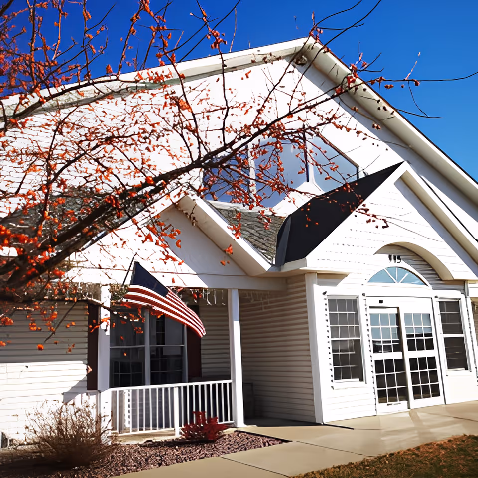 Exterior view of a white building with large windows and a glass door entrance. There is an American flag mounted near the entrance and a tree with red berries in the foreground. The sky is clear and blue.