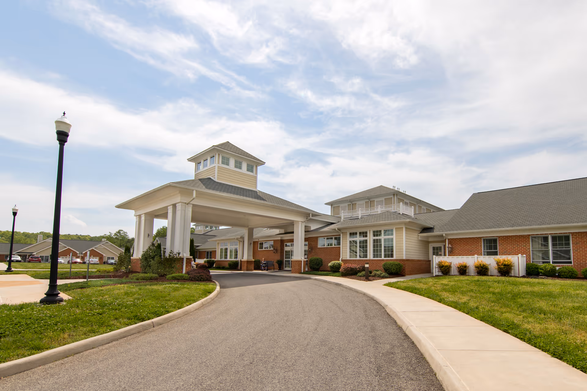 Exterior view of a senior living facility building with a covered entrance driveway, brick and beige siding, large windows, and a well-maintained lawn under a partly cloudy sky.