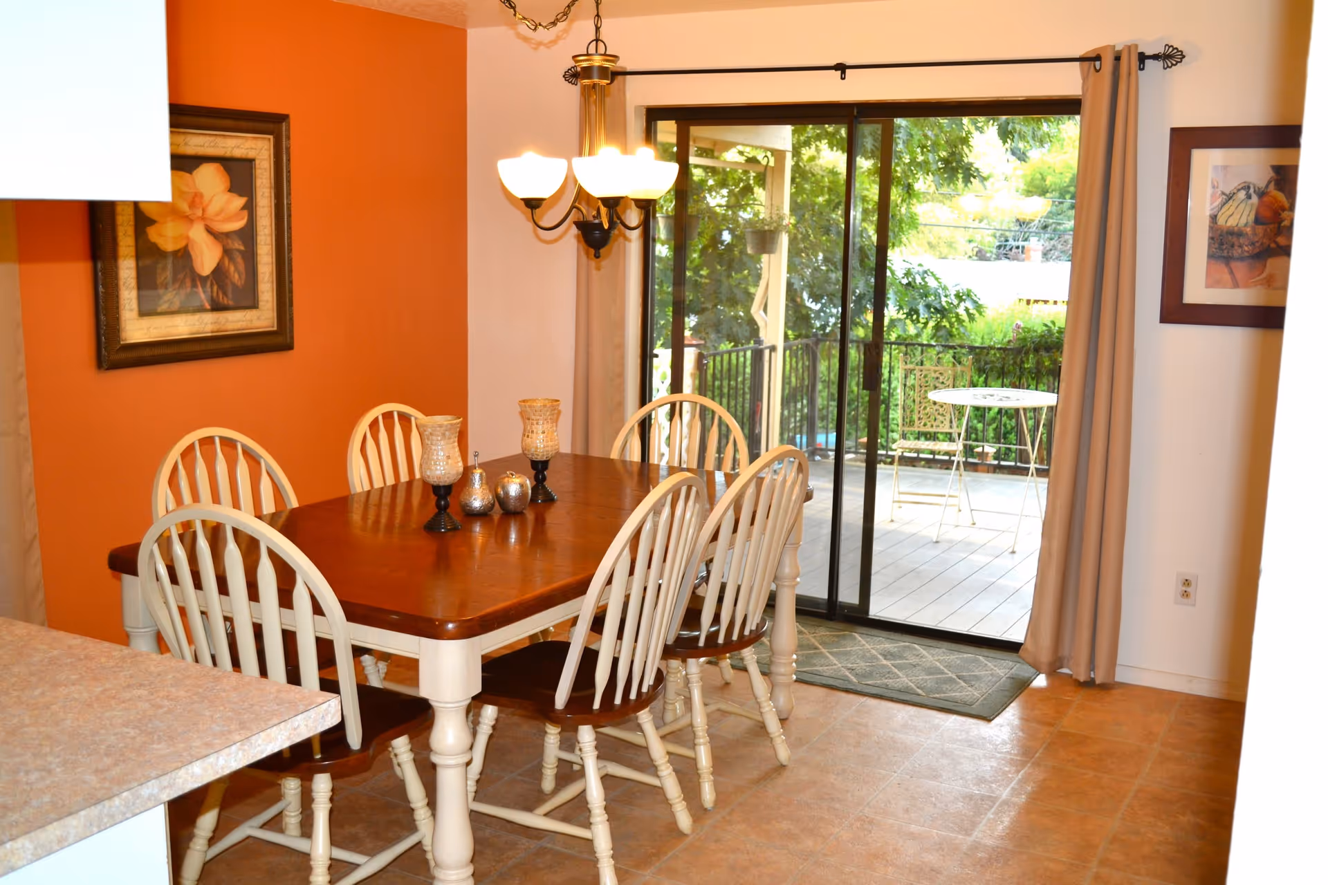 Dining area with a wooden table and six white chairs with dark wooden seats. The table has decorative candle holders and small ornaments. There is a chandelier with five lights hanging above the table. The room has an orange accent wall with a framed flower painting and a sliding glass door with beige curtains leading to an outdoor patio with a small white table and chairs surrounded by greenery.