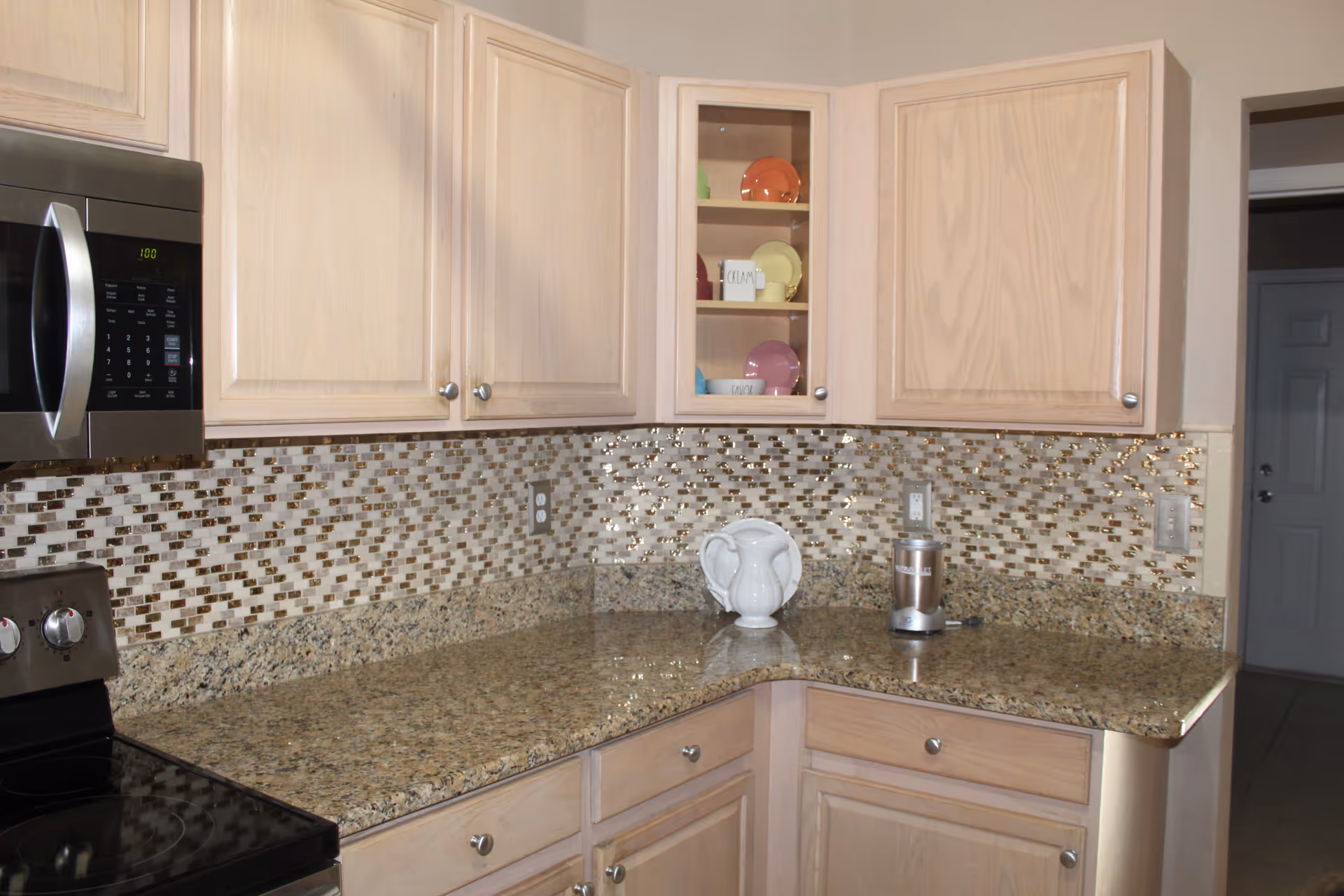 A kitchen corner with light wood cabinets, a granite countertop, and a mosaic tile backsplash in shades of brown and beige. There is a microwave oven mounted above a stove on the left side, a white ceramic pitcher and a small appliance on the countertop, and a cabinet with glass doors displaying colorful plates.