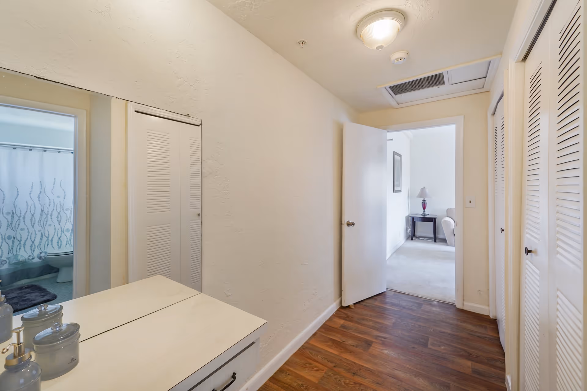 Narrow interior hallway with wood flooring, closets and a dresser with mirror, leading to a living area and a bathroom.
