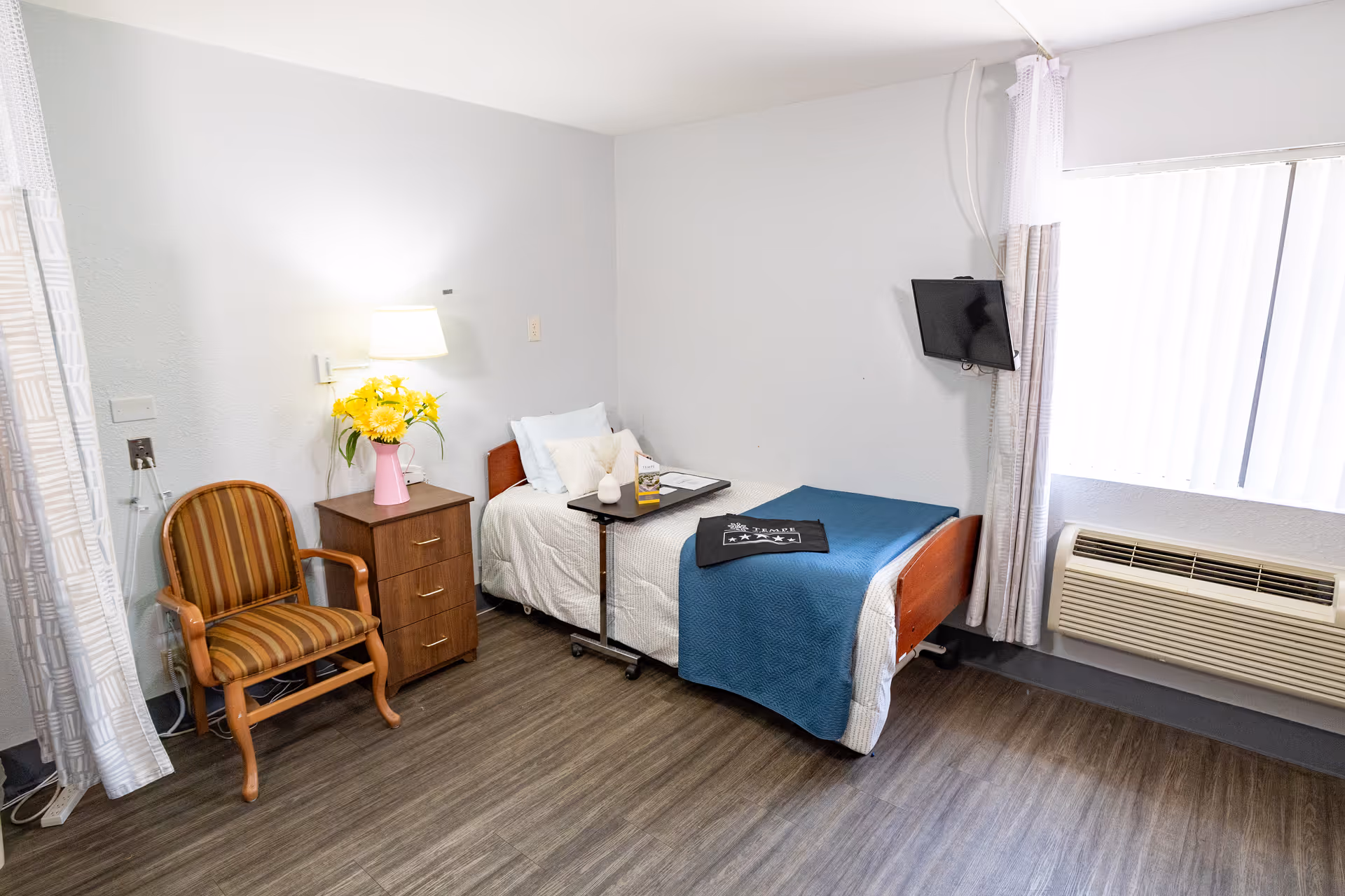 A small, clean bedroom in a senior living facility with a single bed covered with white and blue bedding. Next to the bed is a wooden nightstand with a pink vase holding yellow flowers and a wall-mounted lamp above it. A striped wooden chair is placed beside the nightstand. A flat-screen TV is mounted on the wall near a window with vertical blinds. Below the window is an air conditioning unit. The floor has wood-like vinyl flooring.