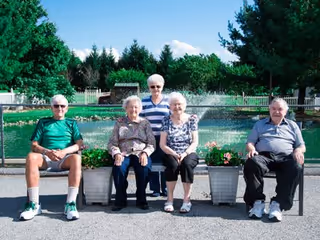 Five elderly individuals sitting on benches outdoors in front of a water fountain with greenery and trees in the background under a clear blue sky.