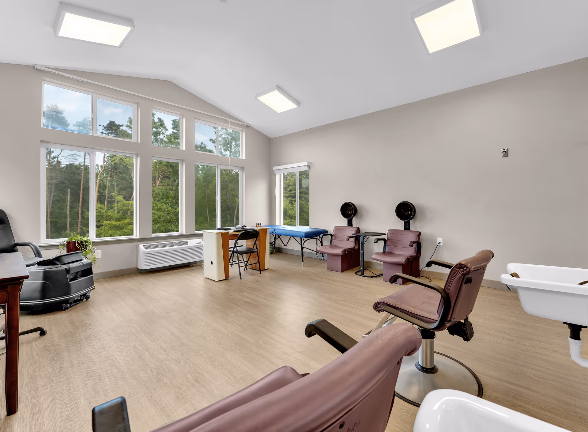 A bright salon room with large windows showing green trees outside. The room has several salon chairs, two hair dryer stations, a manicure table with a chair, and a blue massage or treatment table. The floor is light wood, and the walls are painted a neutral beige color.