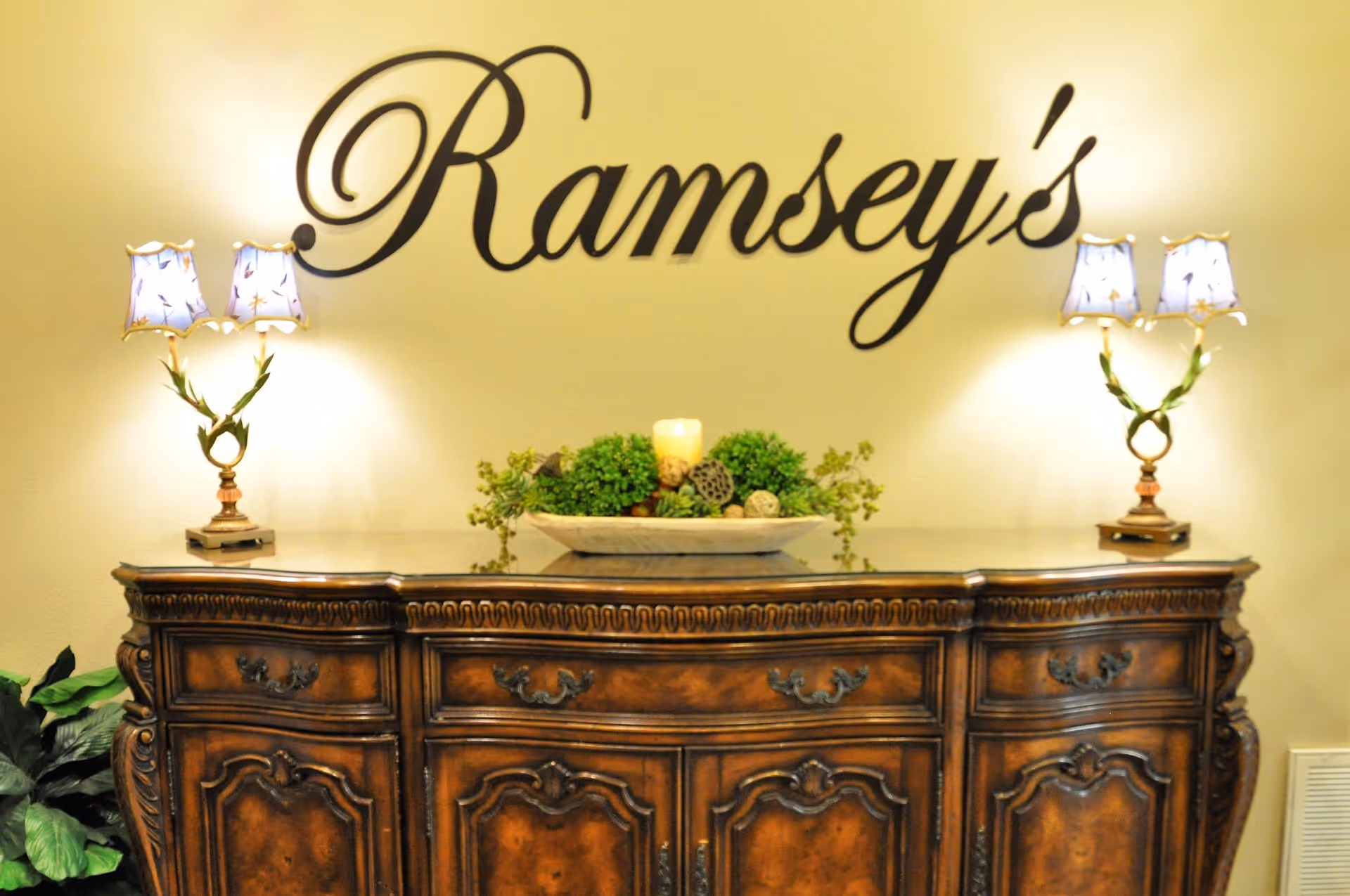 An ornate wooden sideboard with decorative carvings is placed against a beige wall. On top of the sideboard are two matching lamps with floral-patterned shades and a centerpiece featuring greenery and a candle. Above the sideboard, the word 'Ramsey's' is displayed in elegant black cursive lettering on the wall.