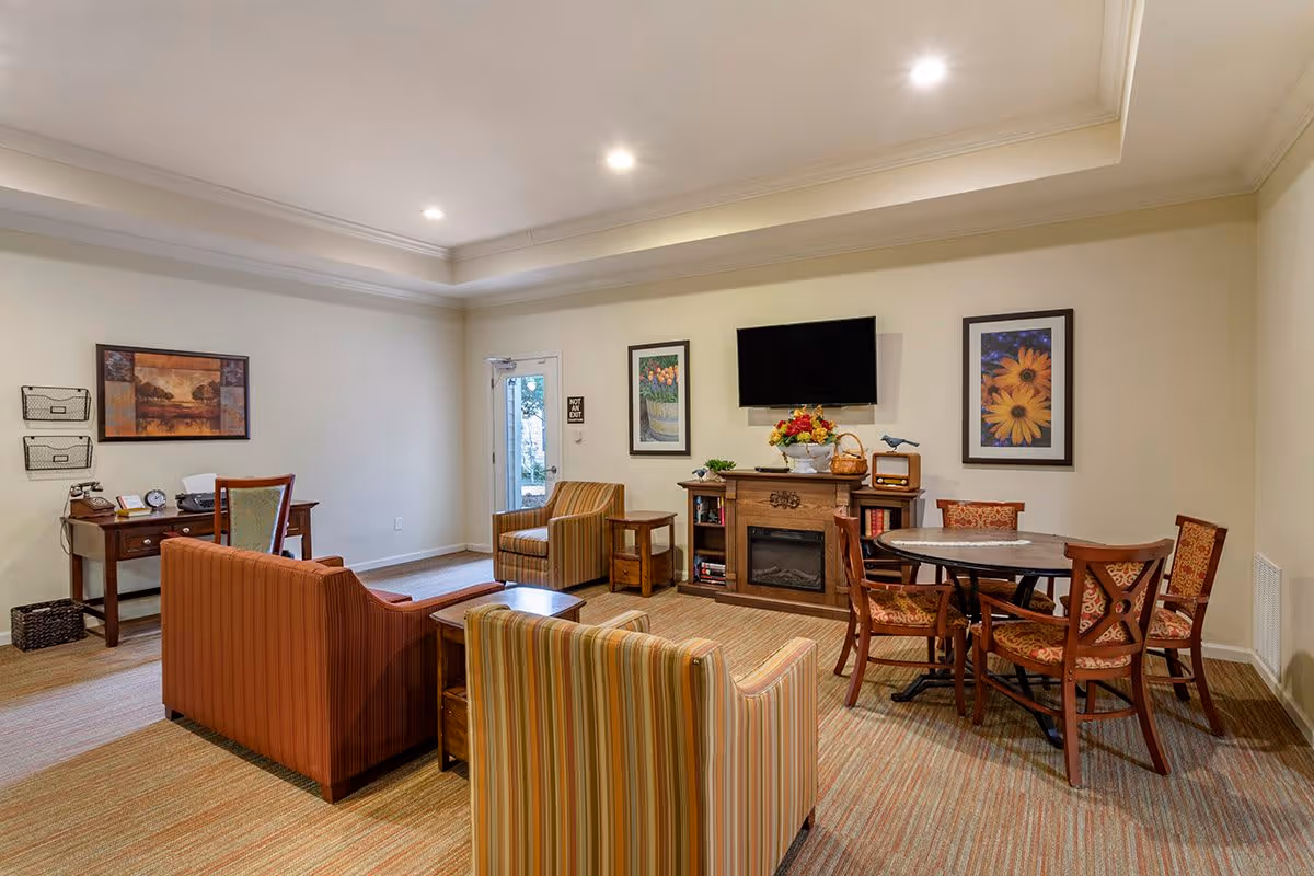 A cozy living room area in an assisted living facility featuring a striped armchair, a brown sofa, a wooden coffee table, and a round dining table with four chairs. The room has a wall-mounted flat-screen TV above a wooden fireplace mantel decorated with a flower arrangement and books. There are framed floral artworks on the walls and a desk with a chair and office supplies in the corner. The room is well-lit with recessed ceiling lights and has a door leading outside.