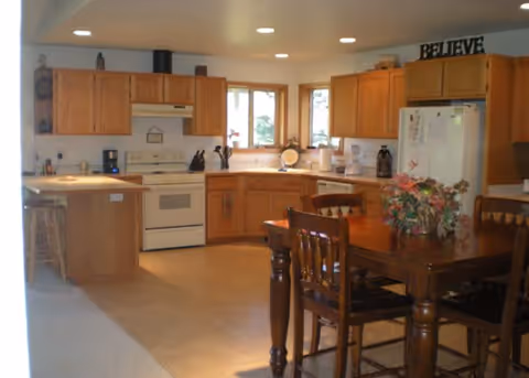 A spacious kitchen with wooden cabinets, a white stove and refrigerator, a coffee maker, and a wooden dining table with chairs. There is a flower arrangement on the table and a decorative sign that says 'BELIEVE' on top of the cabinets.