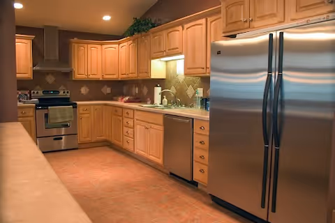 Well-lit kitchen with wooden cabinets, stainless steel refrigerator and stove, and tiled floor.