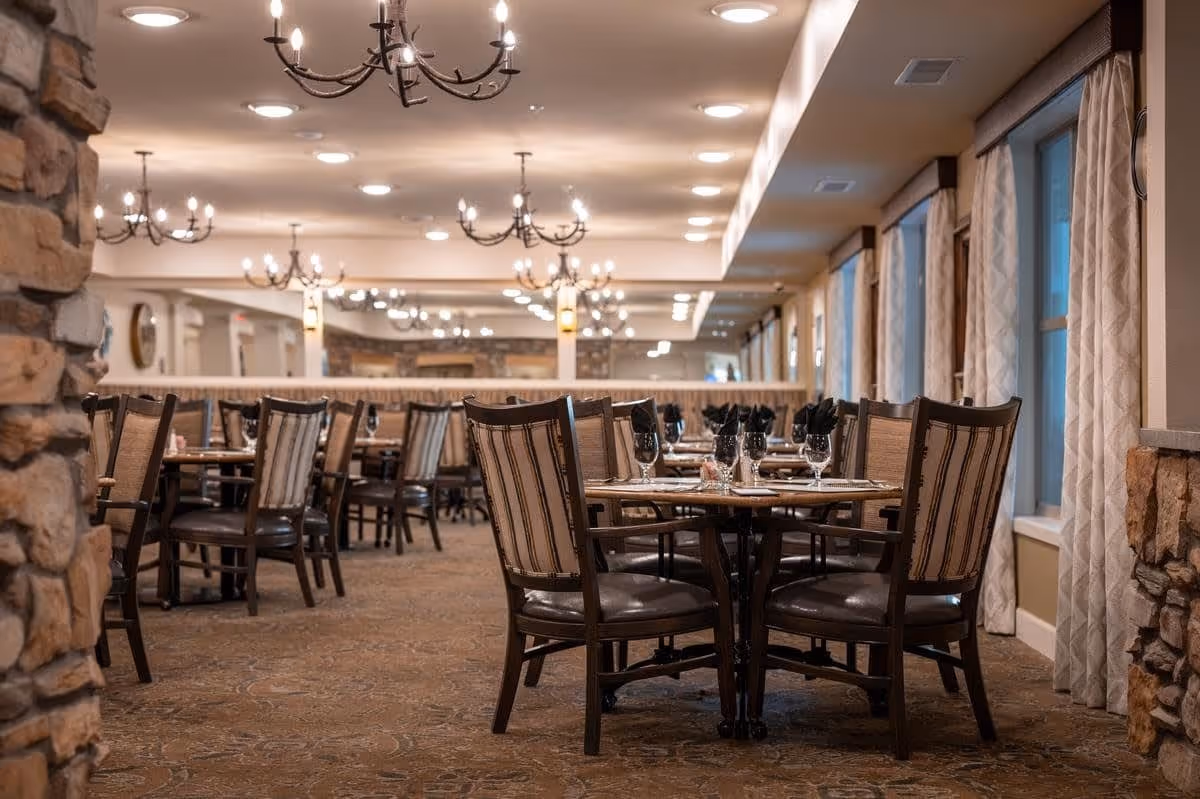 A spacious dining room in a senior living facility with multiple tables and chairs arranged neatly. Each table is set with glassware and black napkins. The room features stone pillars, large windows with curtains, and elegant chandeliers hanging from the ceiling.