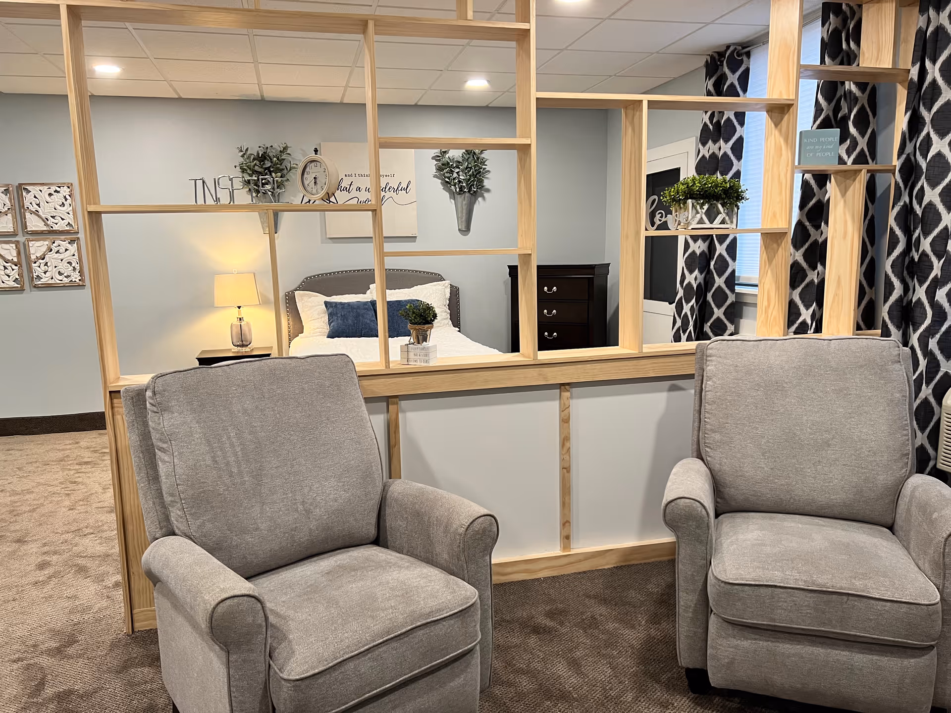 Two gray upholstered armchairs face a wooden open-shelf partition revealing a decorated bedroom with a bed, nightstand, and lamp.