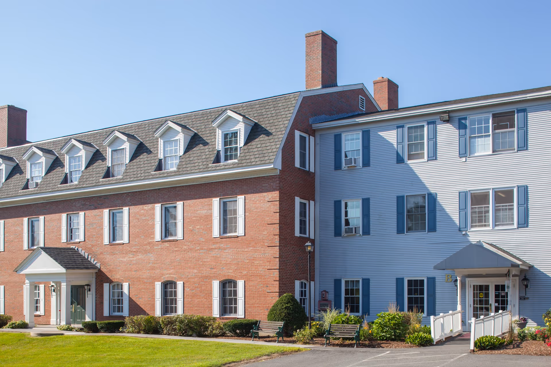 Exterior view of a senior living community building with a combination of red brick and light blue siding. The building has multiple windows with white and blue shutters, two entrances with small porticos, and a well-maintained lawn with shrubs and benches in front.