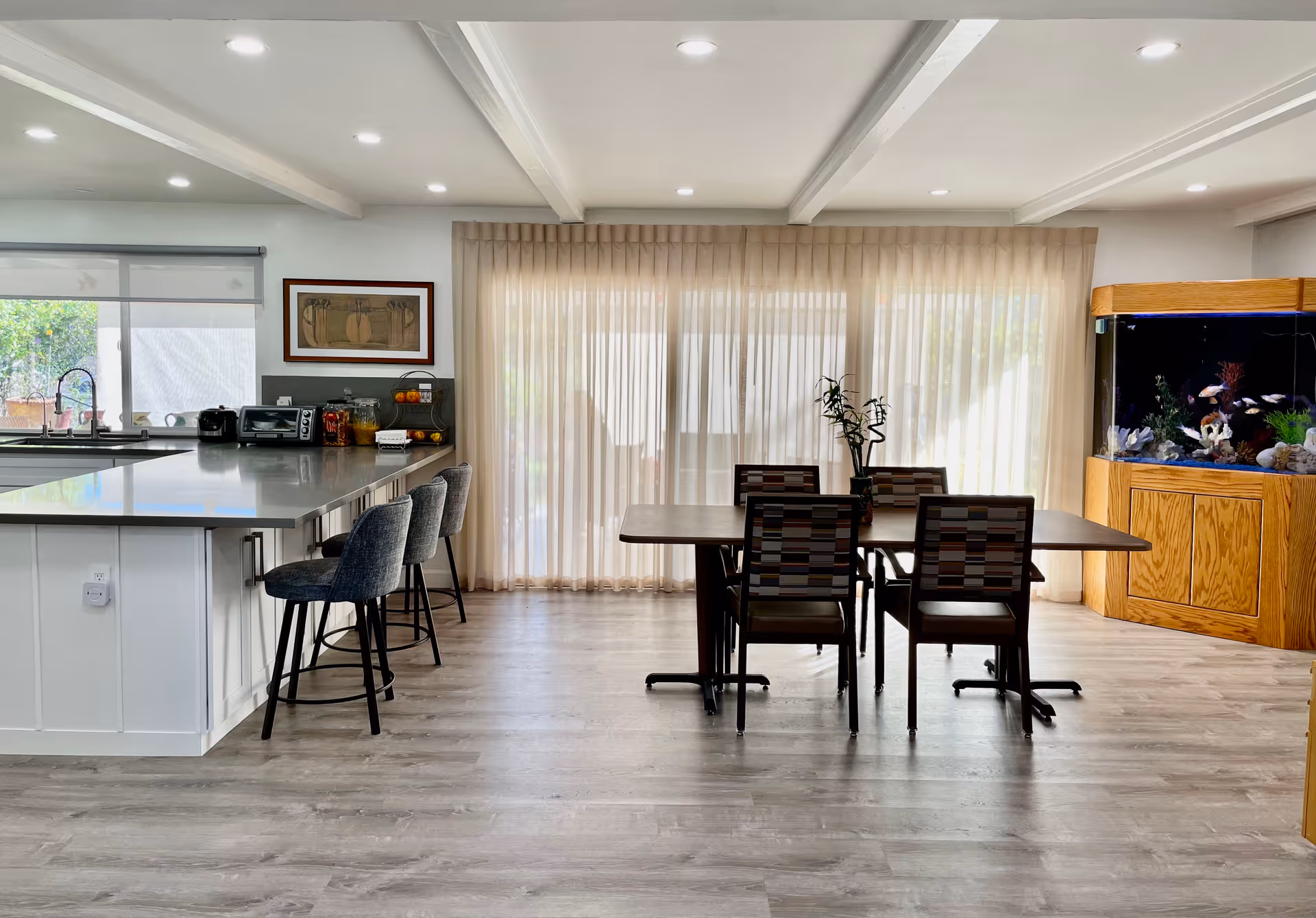 A bright and modern dining area with a rectangular wooden table and four chairs with patterned upholstery. To the left is a kitchen island with a gray countertop and three gray bar stools. The kitchen area includes a sink, toaster oven, and various kitchen items. Large sheer curtains cover the windows in the background, allowing natural light to fill the room. On the right side, there is a large wooden aquarium filled with fish and aquatic plants. The floor is light-colored wood, and the ceiling has recessed lighting.