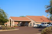 Exterior view of South Mountain Post Acute facility showing a single-story building with a covered entrance, several parked vehicles, and clear blue sky.