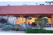 Exterior view of a building with a red roof and wooden porch structure. The porch has hanging flower baskets and outdoor seating with a table and chairs. There are shrubs and plants in the landscaped area in front of the porch.