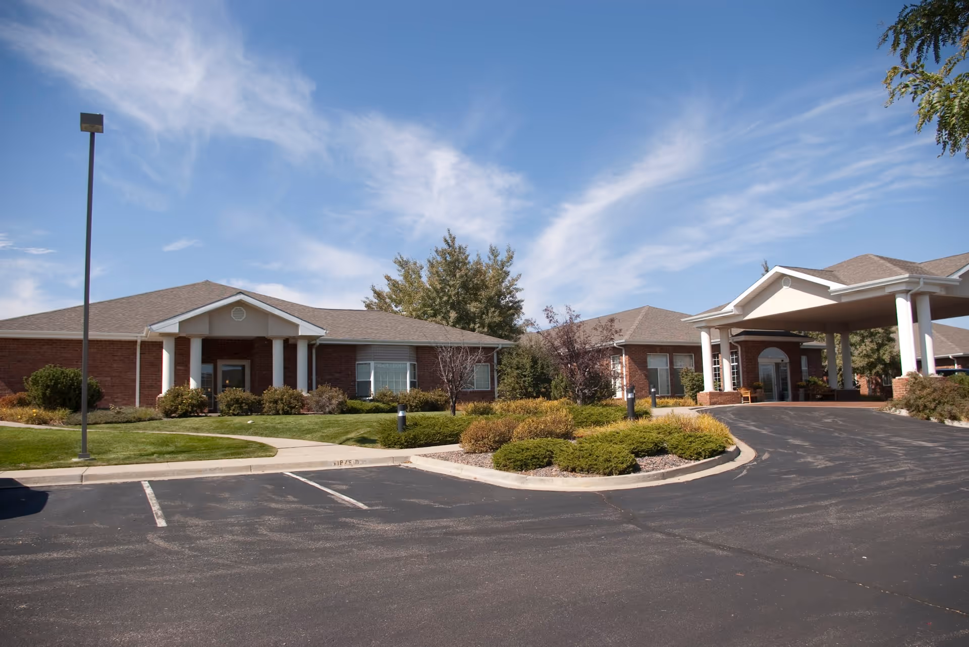Exterior view of a single-story brick building with a covered entrance and columns, surrounded by landscaped bushes and trees under a blue sky with scattered clouds. There is a parking lot in the foreground.