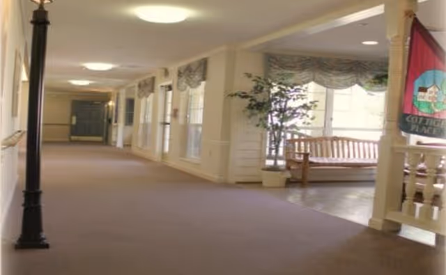 Long carpeted interior hallway with windows, bench seating, decorative plants and a flag in a senior living facility.
