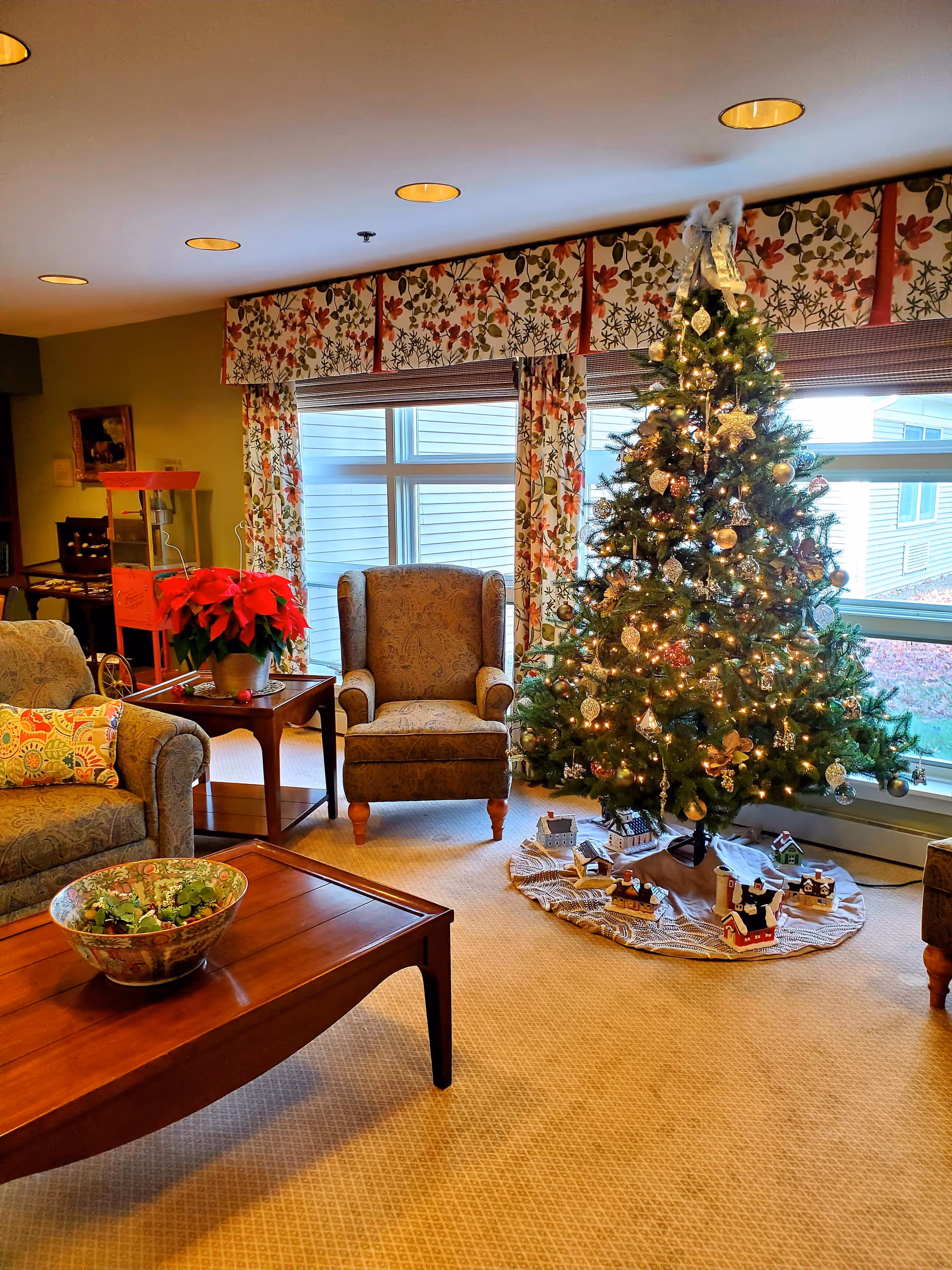 A cozy sitting room with a decorated Christmas tree, two upholstered armchairs, a wooden coffee table and festive decorations by large windows.
