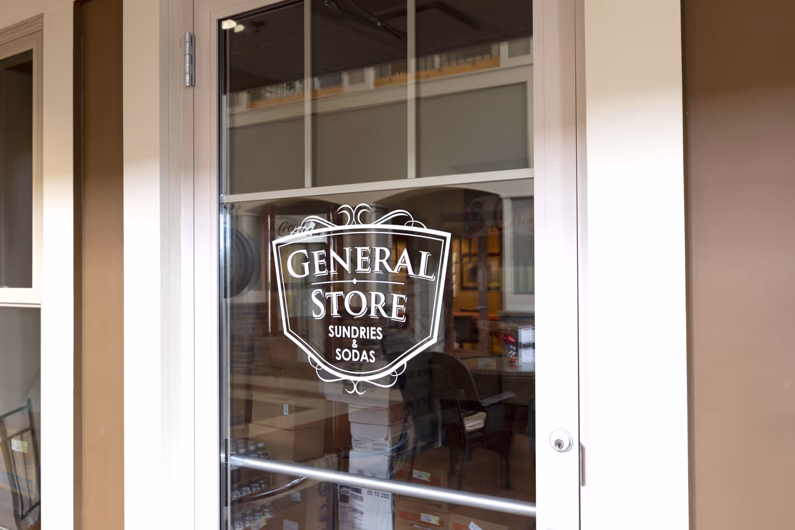 Glass door with a white decal that reads 'GENERAL STORE SUNDRIES & SODAS'. Through the glass, shelves and boxes inside the store are visible along with a chair and some interior lighting.