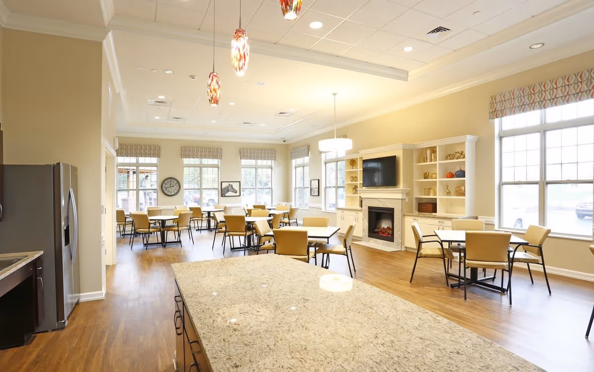 Bright communal dining and lounge area with multiple tables and chairs, a fireplace, and a kitchen island.