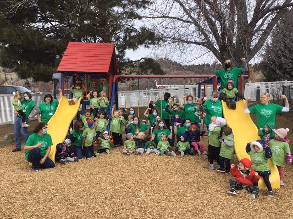 A large group of children and adults wearing matching green shirts gathered around a playground with two yellow slides and a red-roofed play structure. The scene is outdoors with trees and a white fence in the background. Some adults and children are posing with raised arms, and many children are sitting or standing on the wood chip ground.
