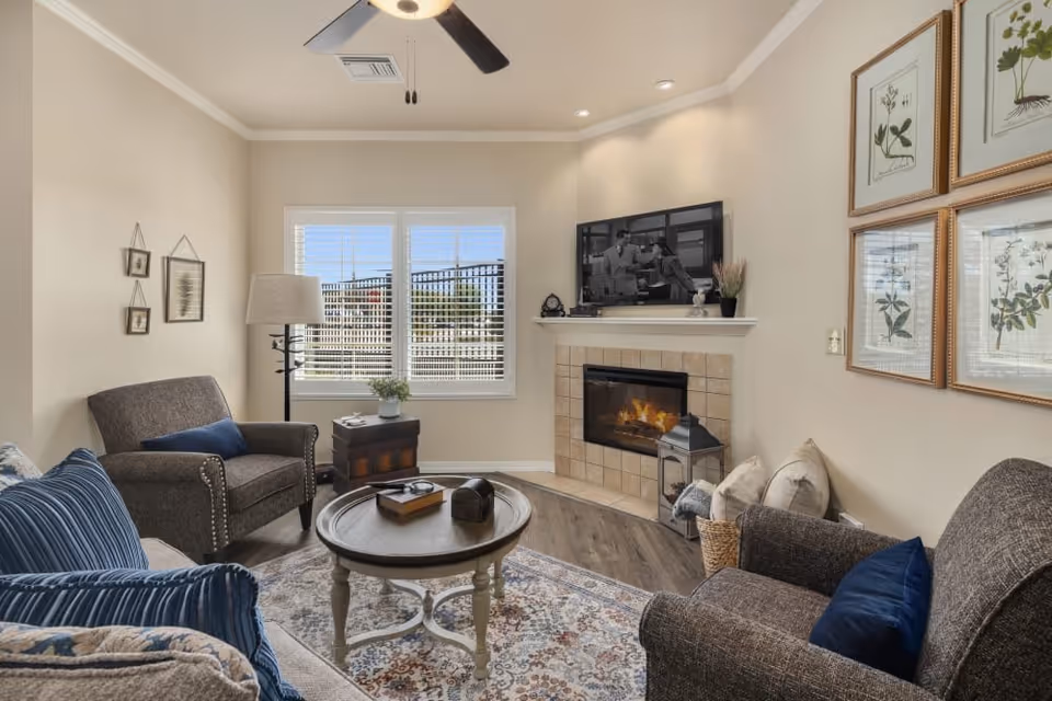 Cozy living room with two armchairs, a round coffee table, a fireplace with TV above, and a window with blinds.