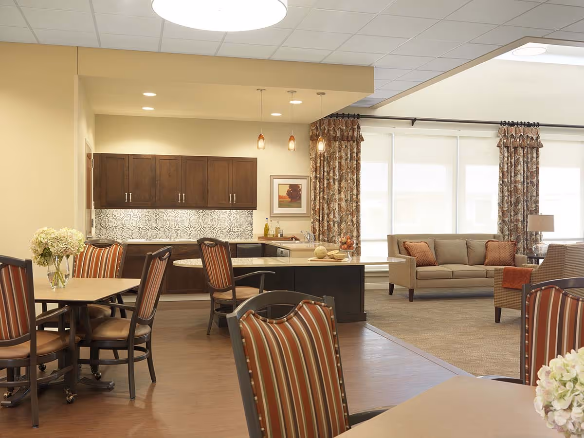 Open communal dining and living area with dining tables and chairs, a kitchen island, sofa and large windows.