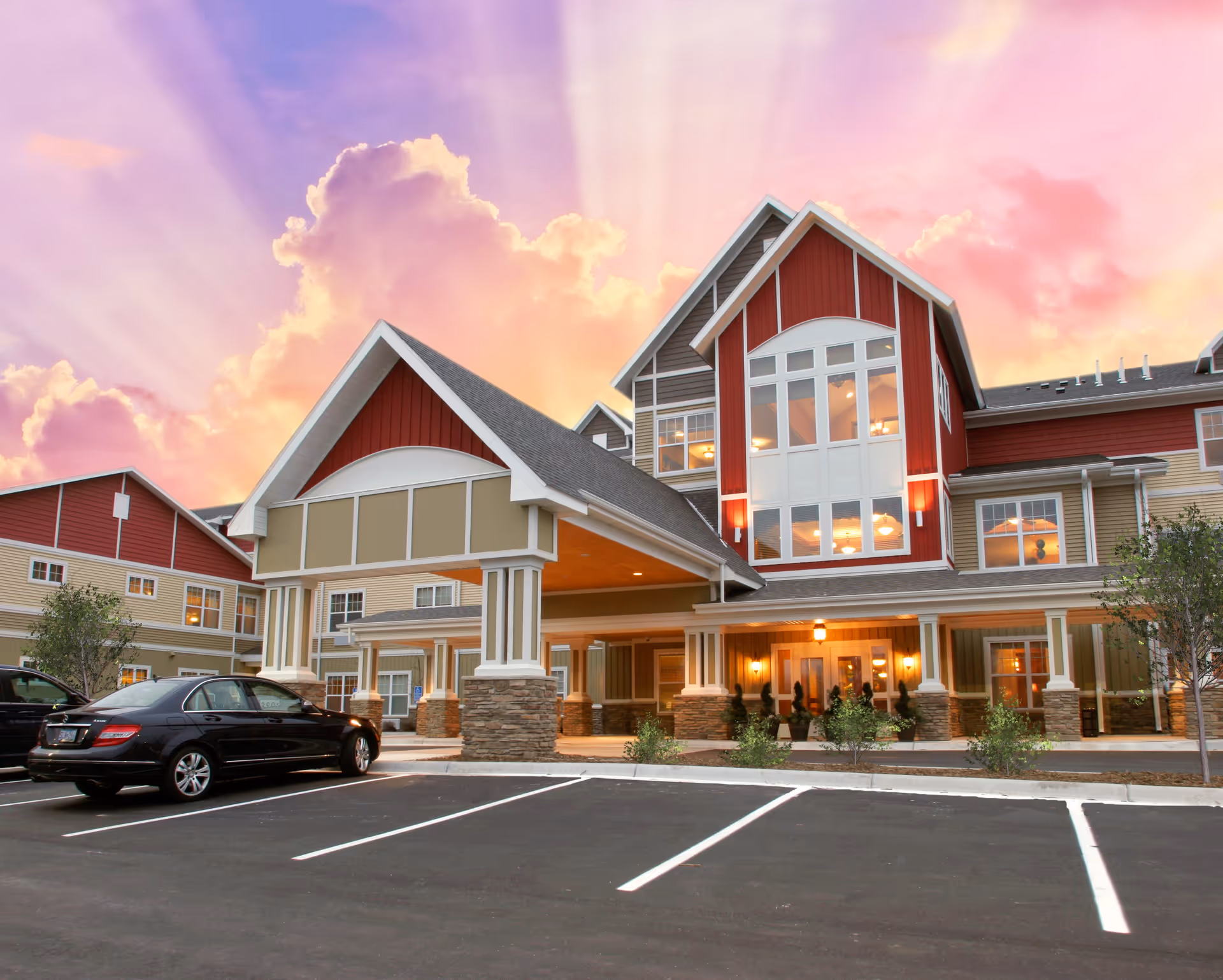 Exterior view of a senior living facility building with a covered entrance, large windows, and a parking lot with a black car parked. The sky is colorful with pink and purple clouds at sunset or sunrise.