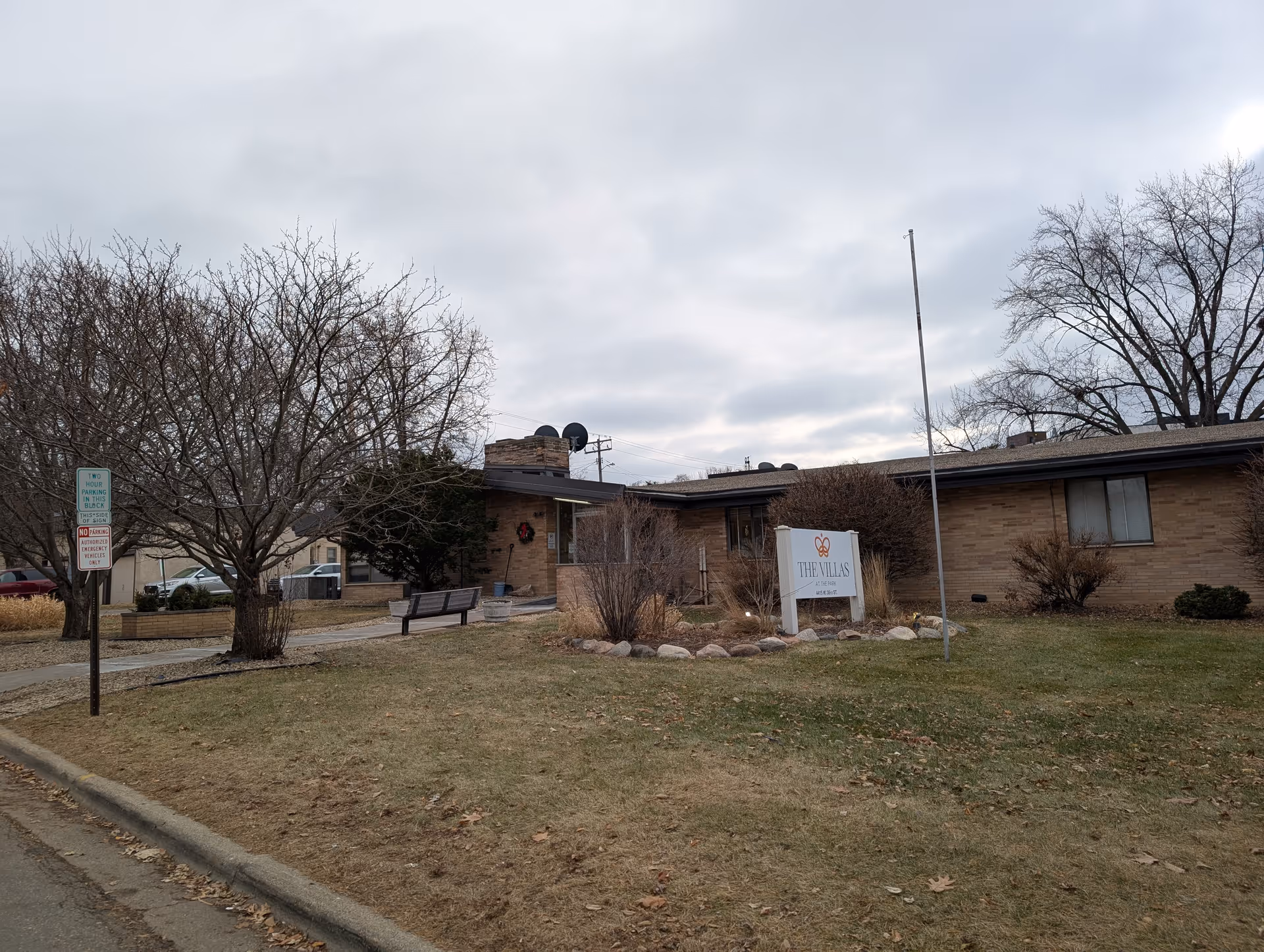 Exterior view of a single-story brick building with a sign in front that reads 'THE VILLAS at Park Health A Villa Center'. The building is surrounded by leafless trees and a grassy lawn under a cloudy sky.