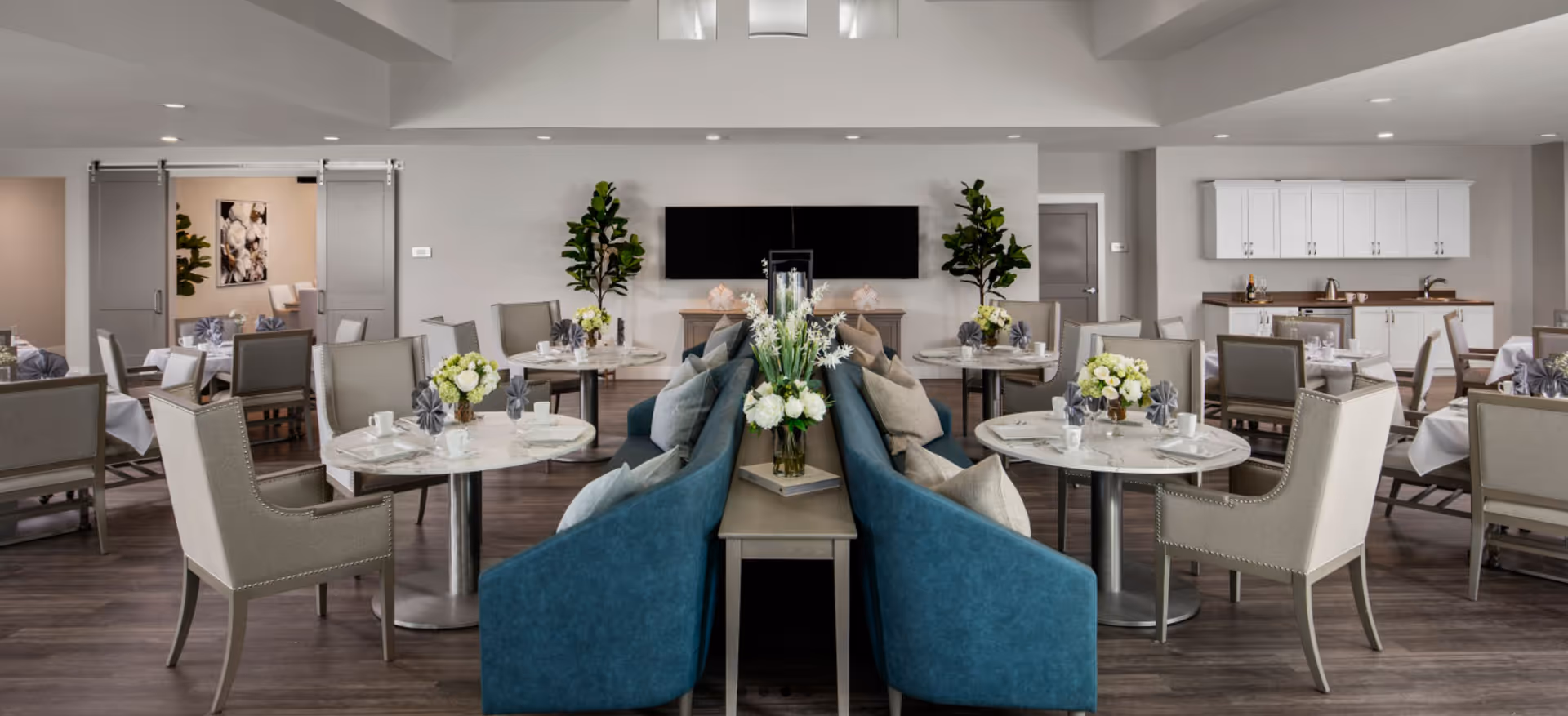 A spacious dining room in a senior living facility with round tables set with white tablecloths, cups, and napkins. The room features comfortable beige chairs and blue cushioned bench seating with pillows. There are flower arrangements on the tables and a flat-screen TV mounted on the wall. The room has wood flooring, white cabinetry with a countertop and sink, and sliding doors leading to another room.