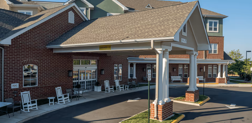 Entrance of a senior living facility with a covered drop-off area supported by white columns on brick bases. The building has red brick walls and multiple windows, with white rocking chairs and small tables placed along the sidewalk near the entrance. The address number 8350 is visible on the covered entrance roof.