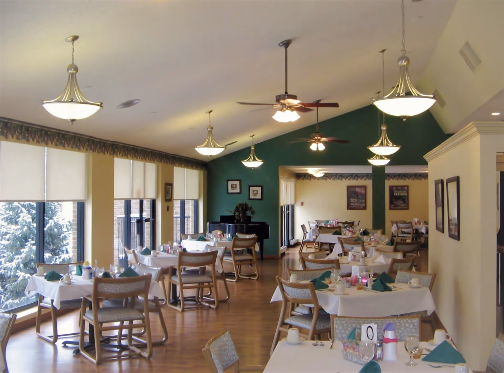 A dining room in Hillside Terrace Senior Living with multiple tables covered in white tablecloths, set with green napkins, glasses, and cups. The room has large windows with blinds, wooden chairs, ceiling fans, and hanging light fixtures. There is a piano against a green accent wall and framed pictures on the walls.