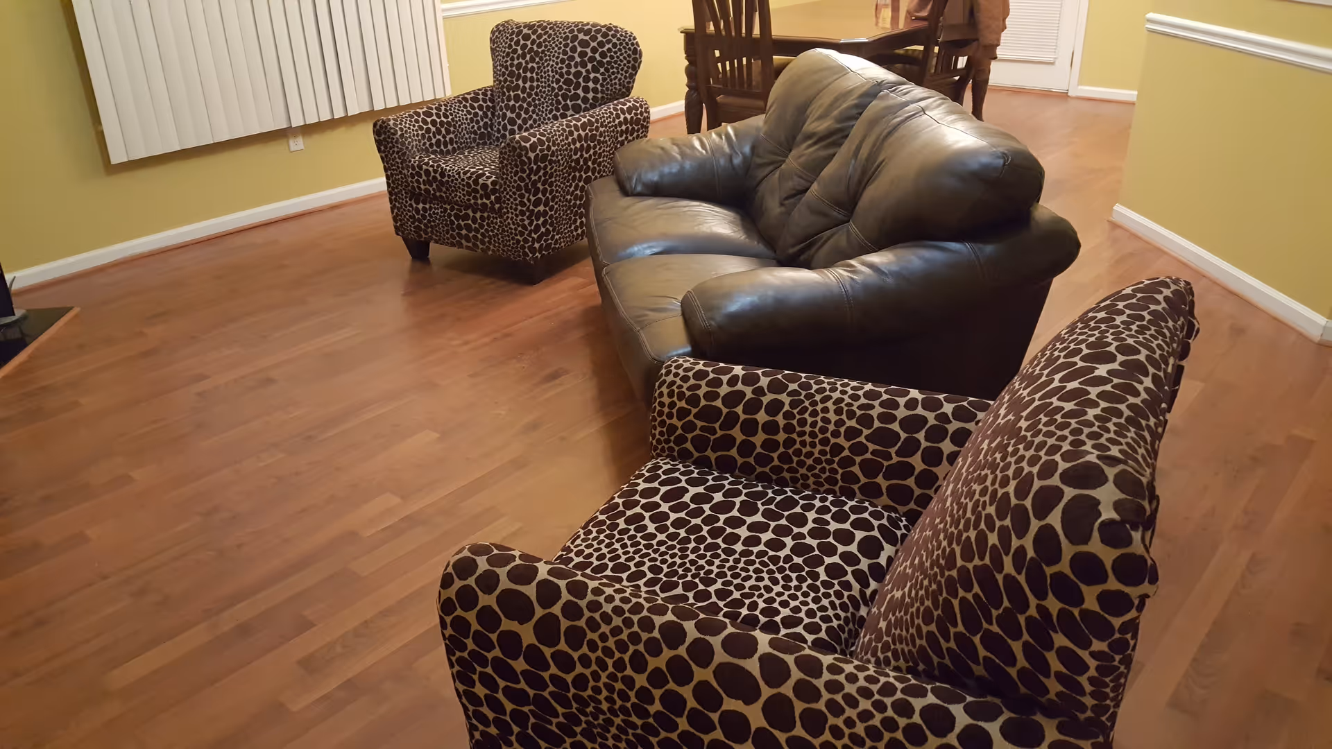 A living room with wooden flooring featuring a black leather couch and two armchairs with a brown and beige spotted pattern. In the background, there is a wooden dining table with chairs and a door with a window blind.