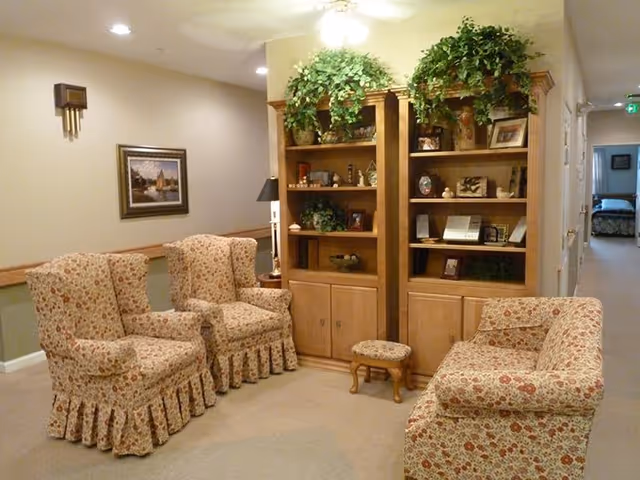 A cozy living room area with two floral-patterned armchairs and a matching sofa arranged around a wooden bookshelf filled with decorative items and plants on top. The room has beige walls, carpeted floor, a framed painting on the wall, and a hallway leading to another room in the background.