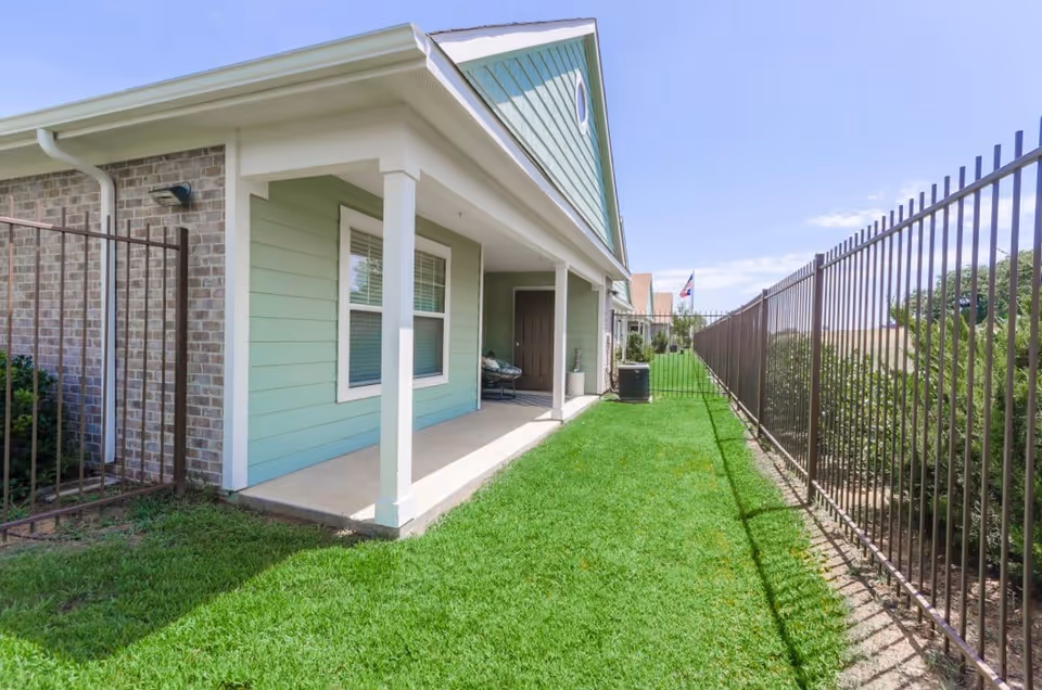 A side view of a residential building with light green siding and white trim, featuring a covered porch area with two white columns. The porch has a chair and a door leading inside. There is a well-maintained green lawn bordered by a metal fence on the right side, and a brick wall on the left side. The sky is clear and blue.