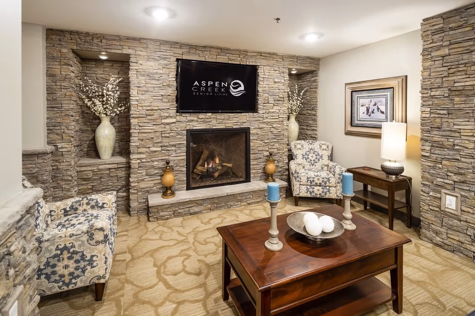 Cozy senior living room with stone walls, a fireplace with a fire burning, a mounted TV displaying the Aspen Creek Senior Living logo, two patterned armchairs, a wooden coffee table with decorative candles and a bowl, a side table with a lamp, and floral arrangements in niches on either side of the fireplace.