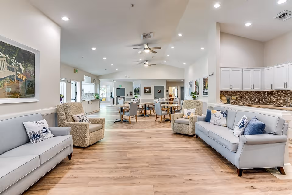 A spacious and well-lit common area in a senior living facility featuring two light gray sofas with decorative pillows, two beige armchairs, wooden flooring, ceiling fans, and a dining area with tables and chairs in the background. The room has white walls adorned with framed artwork and a kitchen area with white cabinets and a mosaic tile backsplash on the right side.