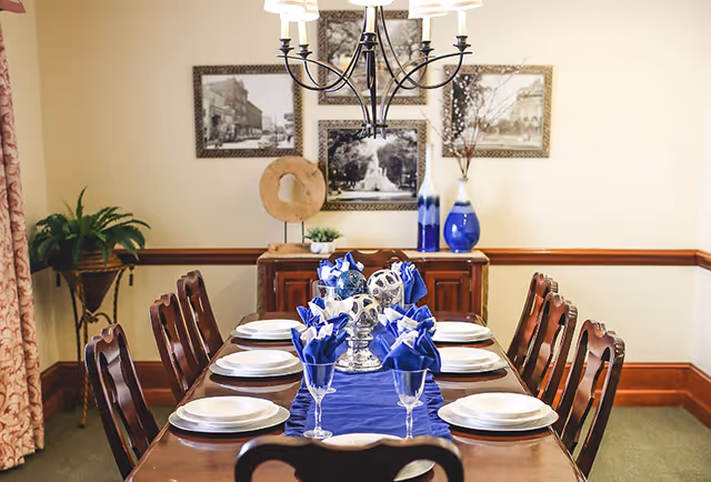 Formal dining room with a long wooden table set with plates, blue napkins, glassware, a chandelier, and a decorative sideboard.