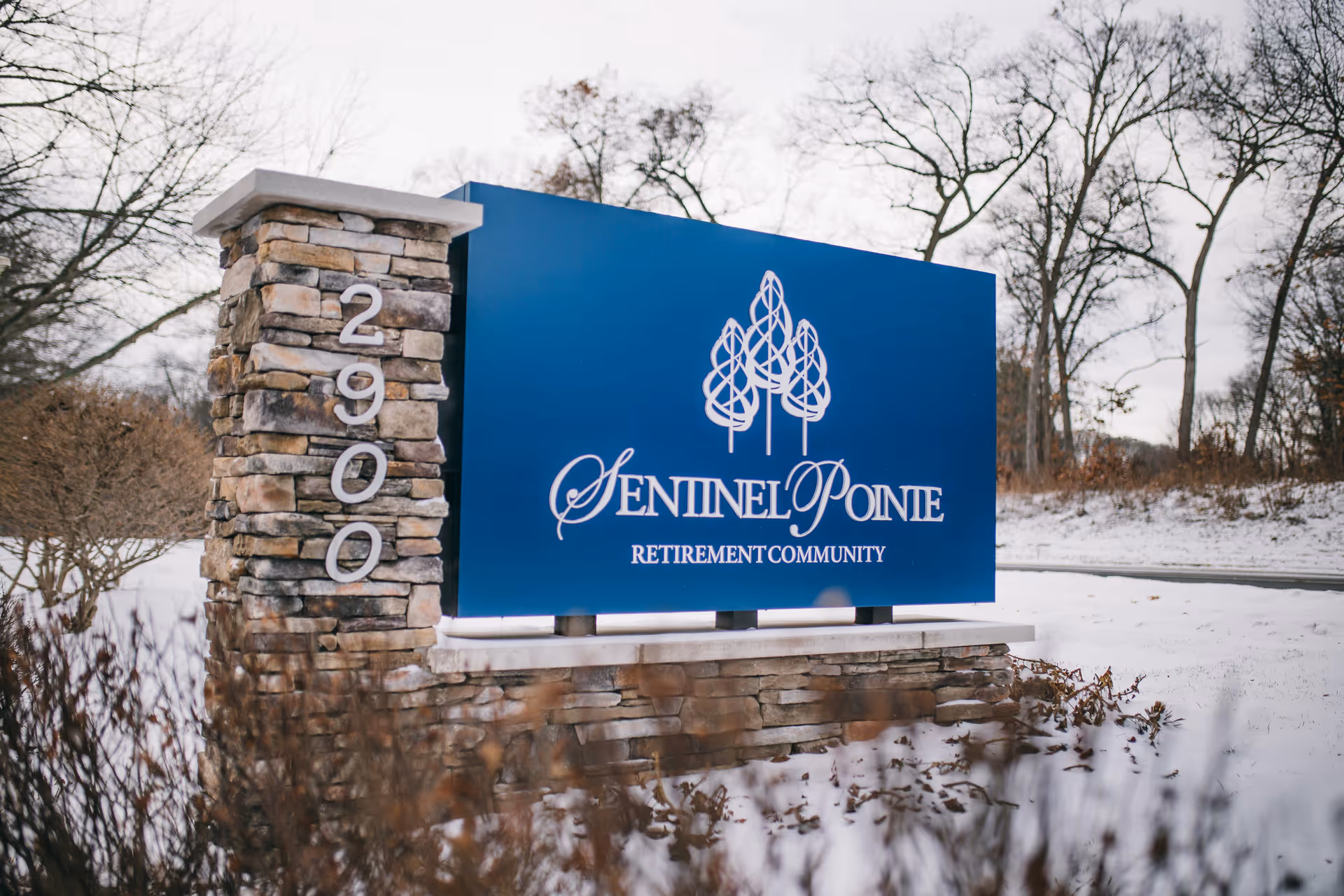 A large blue sign for Sentinel Pointe Retirement Community mounted on a stone base with the address number 2900 displayed vertically on a stone pillar. The surrounding area is covered in snow with leafless trees in the background.