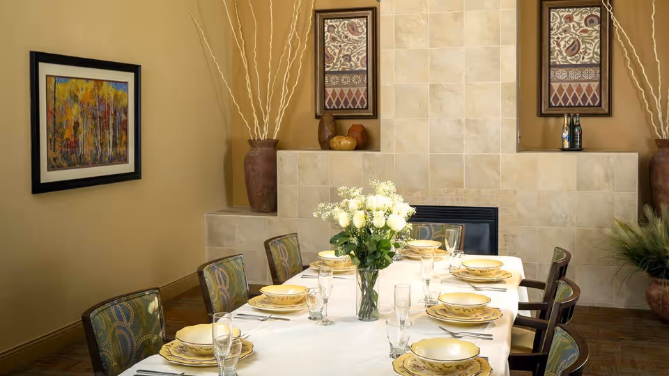 A dining room with a rectangular table set for six people. The table is covered with a white tablecloth and has yellow patterned plates, bowls, and glasses arranged neatly. A vase with white flowers is placed in the center of the table. The room features a tiled fireplace in the background, two framed artworks on the wall, and large decorative vases with tall branches on either side of the fireplace.