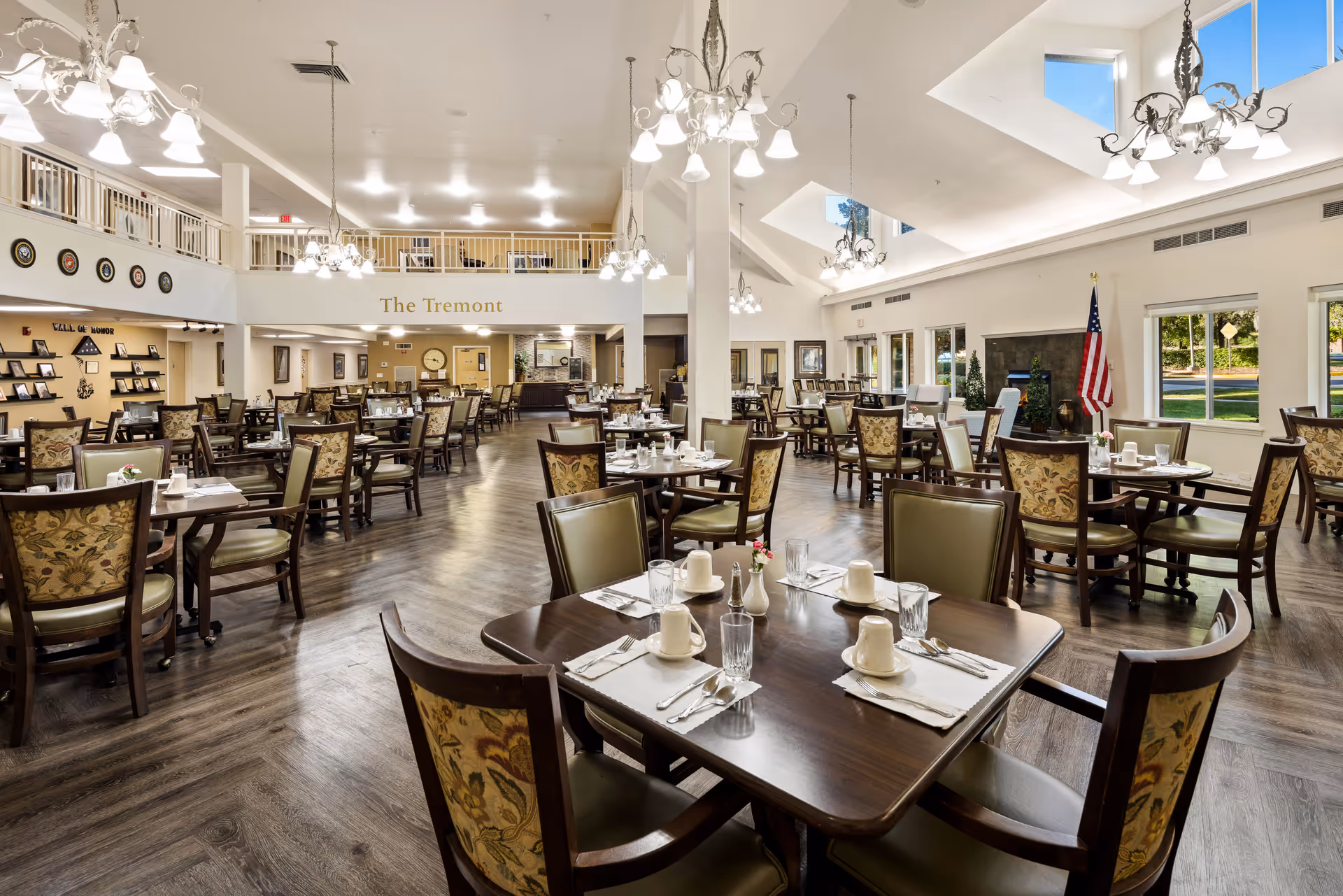 A spacious dining room in The Tremont senior living community with multiple tables set with cups, glasses, and silverware. The room features high ceilings with chandeliers, large windows allowing natural light, and an American flag near a fireplace. The decor includes patterned chairs and wood flooring.