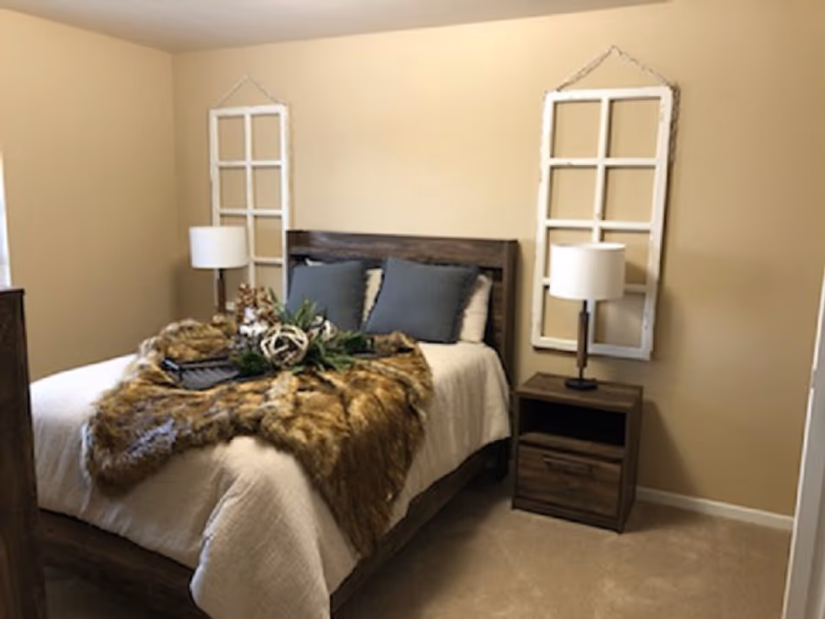 A cozy bedroom with a wooden bed frame and a white bedspread, topped with a brown fur throw and two gray pillows. There are two matching wooden nightstands on either side of the bed, each with a white lampshade. Two decorative white window frame wall hangings are mounted on the beige walls above the nightstands.