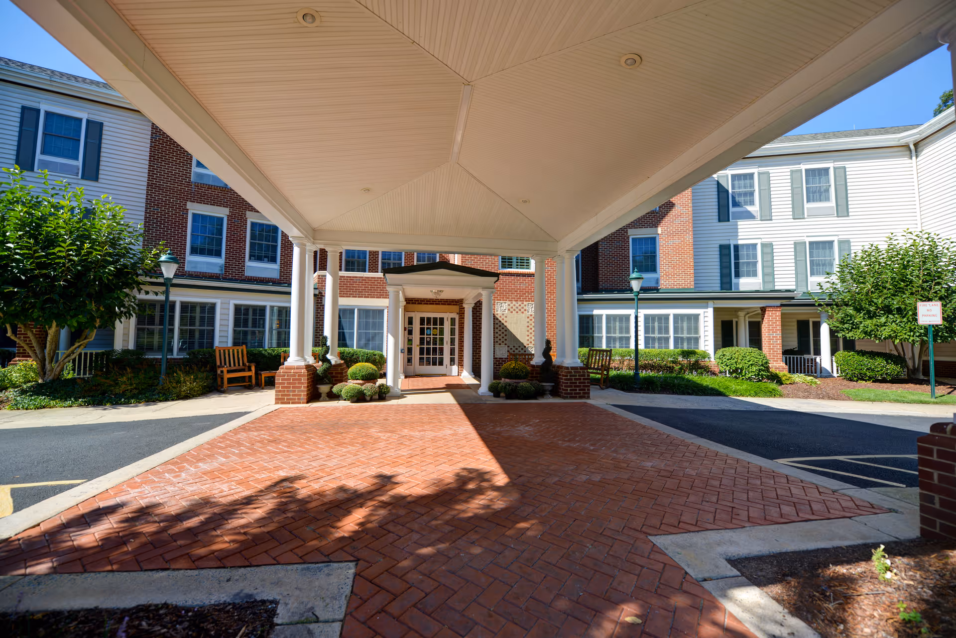 View of the covered entrance area of a senior living facility with brick pavement, white pillars, and a building facade featuring red brick and white siding with multiple windows and green shutters. There are benches, bushes, and trees on either side of the entrance.